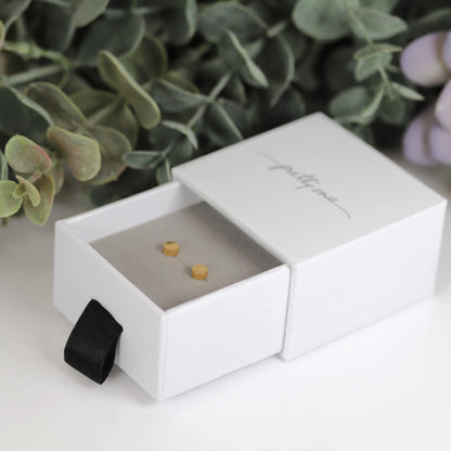 A pair of round dot stud earrings in silver and gold colors, placed on a white surface with foliage in the background.