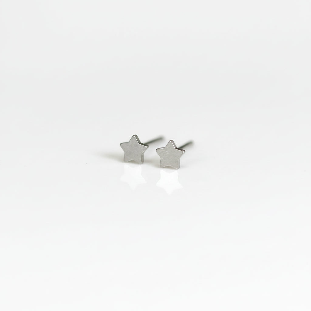 A pair of titanium stud earrings, one shaped like a moon and the other like a star, placed on a reflective surface with a plant in the background.
