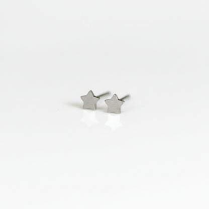 A pair of titanium stud earrings, one shaped like a moon and the other like a star, placed on a reflective surface with a plant in the background.