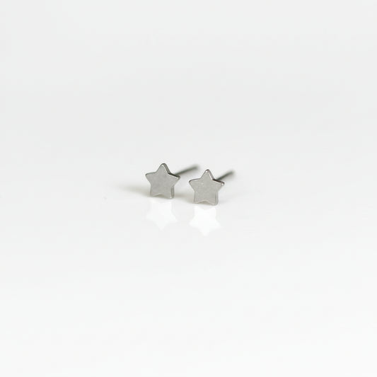A pair of titanium stud earrings, one shaped like a moon and the other like a star, placed on a reflective surface with a plant in the background.