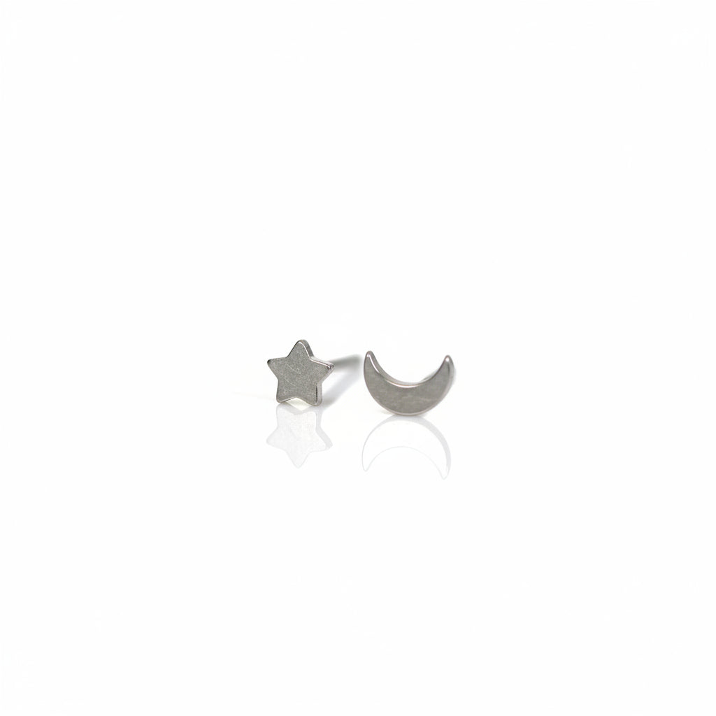 A pair of titanium stud earrings, one shaped like a moon and the other like a star, placed on a reflective surface with a plant in the background.