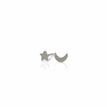 A pair of titanium stud earrings, one shaped like a moon and the other like a star, placed on a reflective surface with a plant in the background.