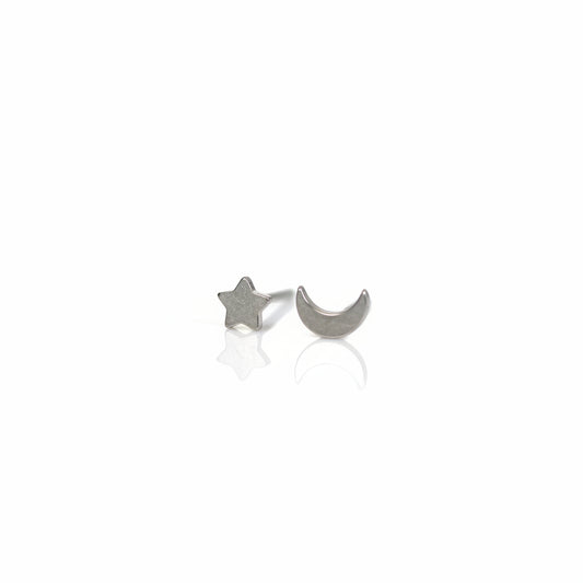 A pair of titanium stud earrings, one shaped like a moon and the other like a star, placed on a reflective surface with a plant in the background.