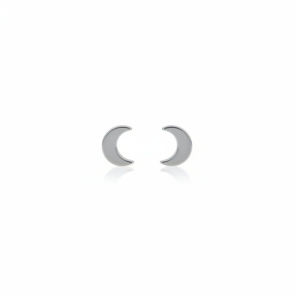 A pair of titanium stud earrings, one shaped like a moon and the other like a star, placed on a reflective surface with a plant in the background.