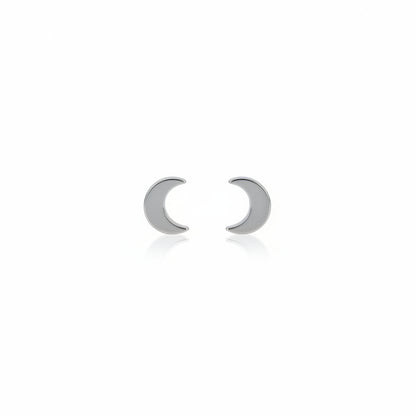 A pair of titanium stud earrings, one shaped like a moon and the other like a star, placed on a reflective surface with a plant in the background.