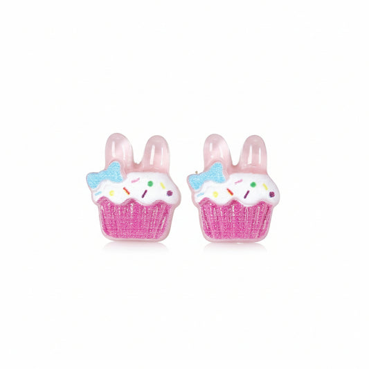 A pair of pink cupcake-shaped stud earrings with bunny ear designs, adorned with white and blue accents, displayed on a reflective surface.