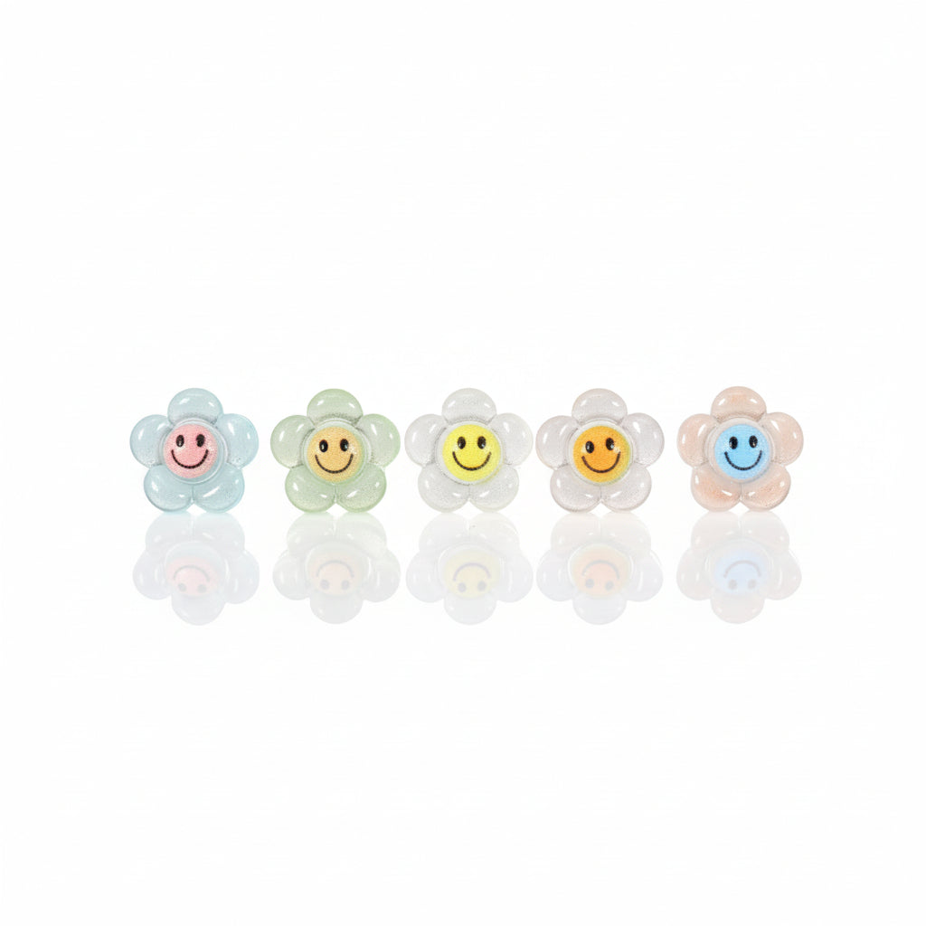A series of glow-in-the-dark smiley face stud earrings in various colors displayed against a white background with green foliage in the backdrop.