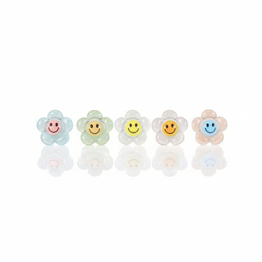 A series of glow-in-the-dark smiley face stud earrings in various colors displayed against a white background with green foliage in the backdrop.
