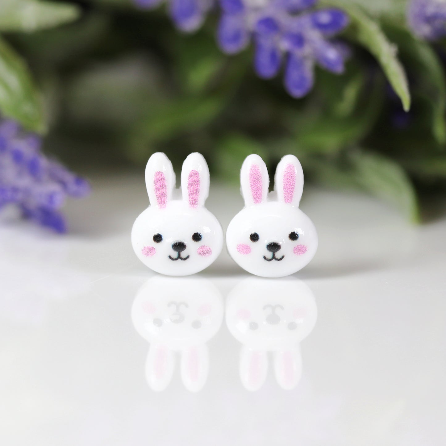 A pair of white bunny face stud earrings with pink inner ears and black eyes, attached to clear posts.