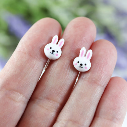 A pair of white bunny face stud earrings with pink inner ears and black eyes, attached to clip on backs
