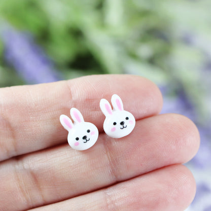 A pair of white bunny face stud earrings with pink inner ears and black eyes, attached to clear posts.