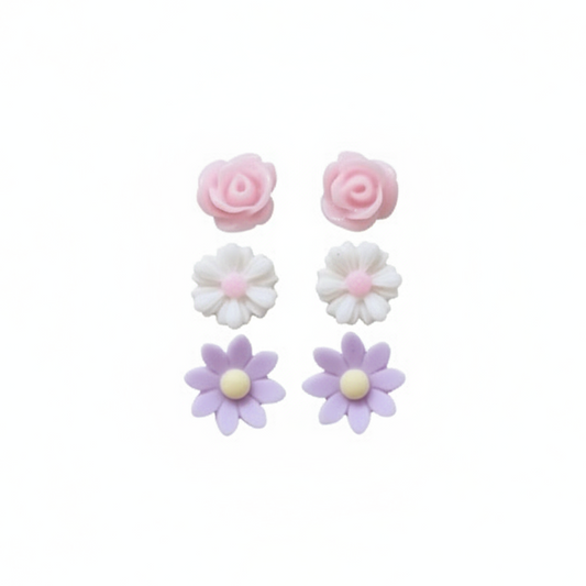 A hand holding three pairs of stud earrings, featuring pink rose, daisy, and wildflower designs in white, pink, and purple colors.