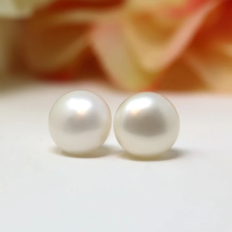 A pair of white, button-style freshwater pearl stud earrings displayed against a soft, floral background.